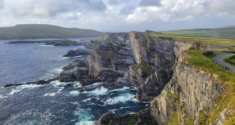Dramatic sea cliffs with layered rock faces meeting turquoise Atlantic waves under a partly cloudy sky.