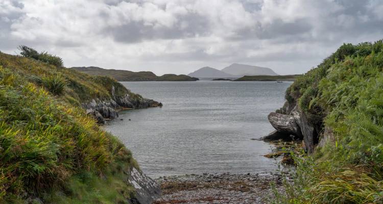 Quiet inlet opening to the sea with distant hazy peaks beneath a grey-cloud sky.