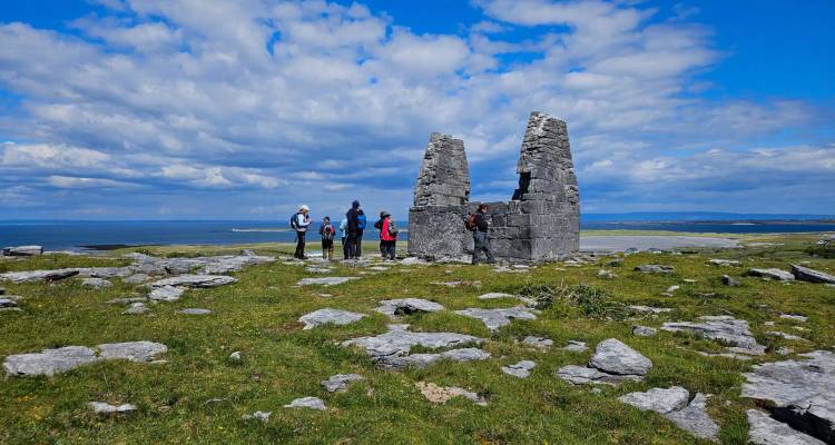 Wandergruppe versammelt sich um eine kleine zerstörte Steinstruktur auf einem windigen Kalksteinplateau mit Meerblick im Hintergrund.