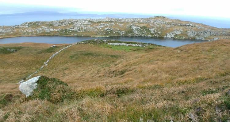 Rolling brown-gold hills surround a small lake on a remote coastal peninsula under an overcast sky.