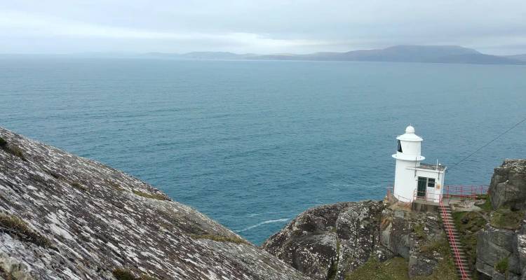 A white lighthouse perched on a steep rocky headland overlooking a vast blue ocean.