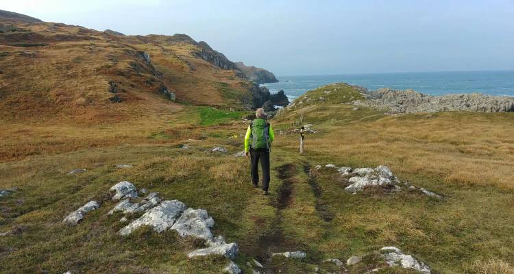 A lone hiker with a green backpack walks along a rugged coastal path with sea views.