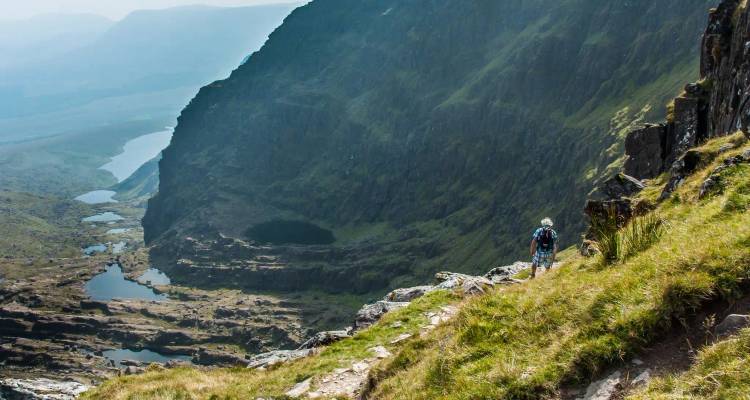 A hiker traverses a steep grassy ridge high above dramatic valley lakes and cliffs.