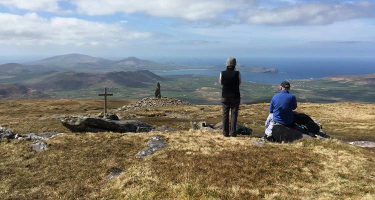 Walkers pause beside a summit cross enjoying panoramic coastal and mountain views.