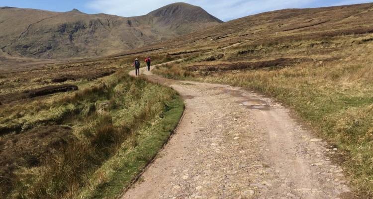 Two walkers follow a rough stony track across open moorland beneath rolling hills.