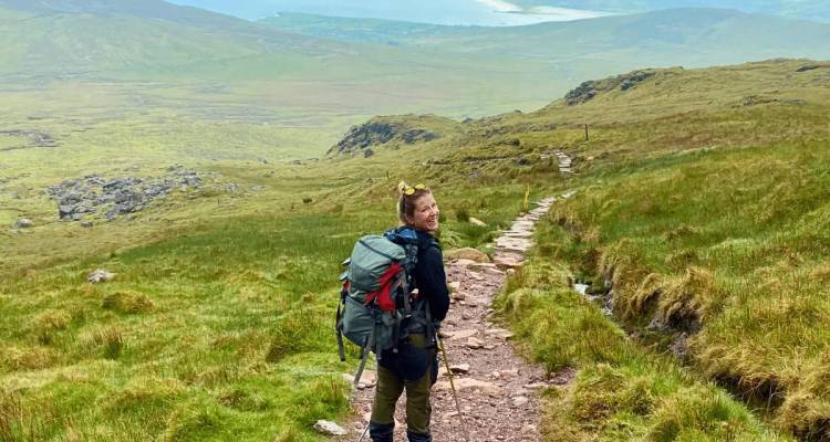 Smiling backpacker climbs rocky mountain trail with wide valley and lakes fading into mist below.