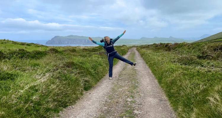 Joyful hiker jumping with arms outstretched on a gravel track through grassy coastal countryside.