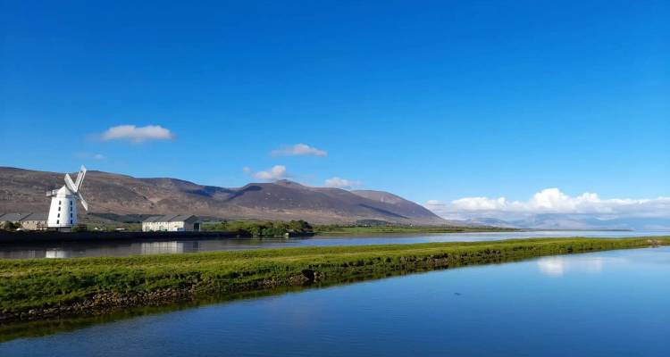 A white traditional windmill stands beside calm water with distant brown mountains under blue sky.
