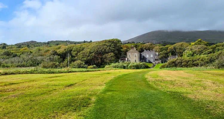 A stately country house nestles among trees at the foot of a mist-capped mountain across open fields.