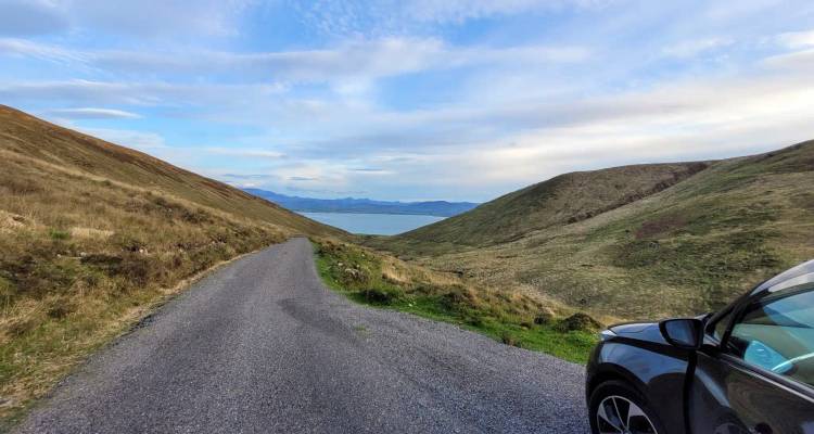 A narrow mountain road descends toward a blue bay, with a parked car at the roadside.