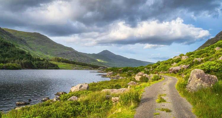 A quiet single-track road winds beside a lake and lush green mountains under moody clouds.
