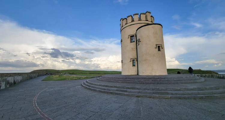 A circular stone lookout tower stands on a raised platform against a blue sky with scattered clouds, surrounded by open green coastal land.