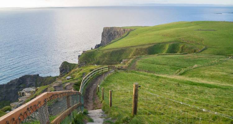 A winding coastal path with railing leads along grassy cliffs overlooking the calm sea.