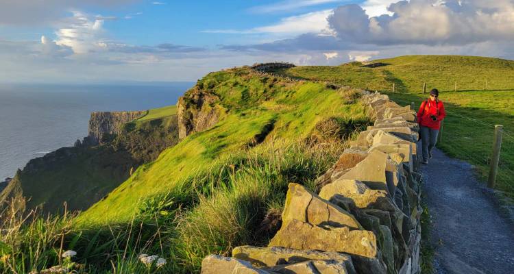 A hiker in a red jacket walks a stone-lined trail along vibrant green cliffs under a bright sky.