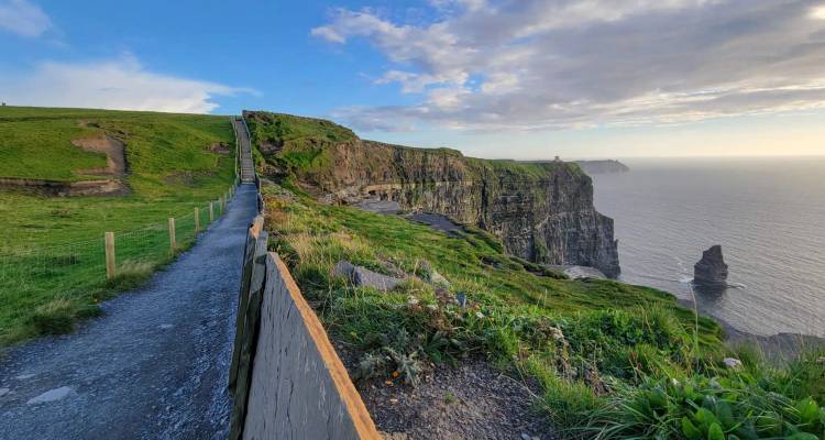 A coastal walking trail bordered by wooden fencing follows rugged cliffs with sea stacks below.