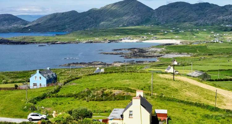 Panoramic view of green fields, scattered cottages and turquoise bay backed by rugged mountains.