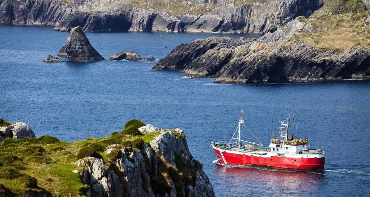 Bright red fishing trawler navigating deep blue water beside rugged rocky stacks.