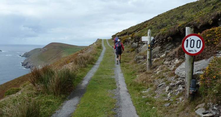 Backpacker walking a narrow grassy track along coastal cliffs past a humorous 100km/h signpost.