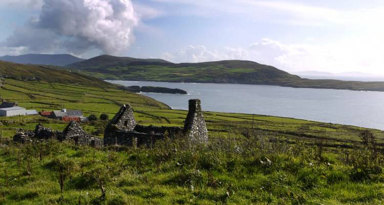 Ruined stone cottage overlooking green patchwork fields and a wide bay under a bright sky.