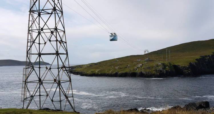 Cable car gliding over choppy water between grassy headlands, supported by a tall metal tower.