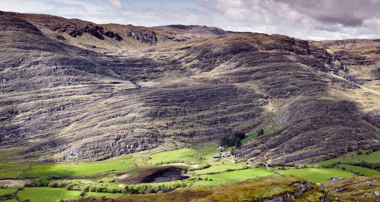 Broad mountain landscape with striated rock slopes overlooking a small lake and lush valley.