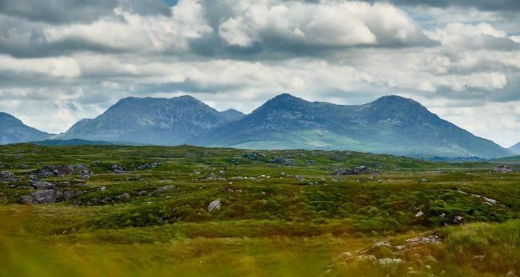 Mountain peaks rise behind a broad green bogland under a sky filled with dramatic clouds.
