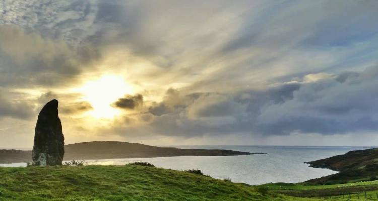 A solitary standing stone silhouetted against a glowing sunset over the sea.