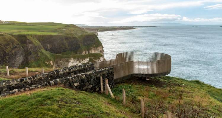 A modern circular wooden viewing platform juts out over high cliffs overlooking the ocean.