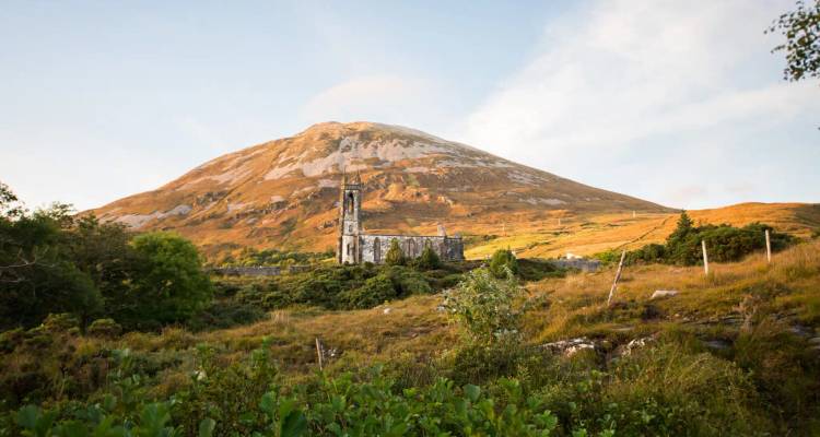 Ruins of a stone church sit below a sunlit mountain during early evening light.
