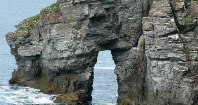 Close-up of a natural sea arch carved into layered grey rock with waves crashing below.