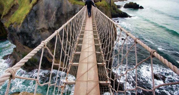 A person crosses a narrow rope bridge suspended high above rugged cliffs and crashing waves.