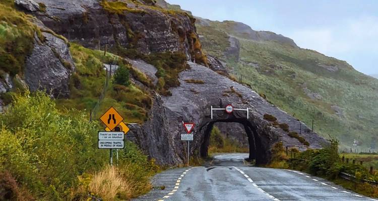 A rural asphalt road curves through a small rock tunnel in a rugged green mountain landscape.
