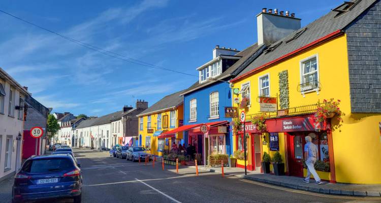 Vibrant brightly painted shops line a sunny Irish village street with café tables outside.