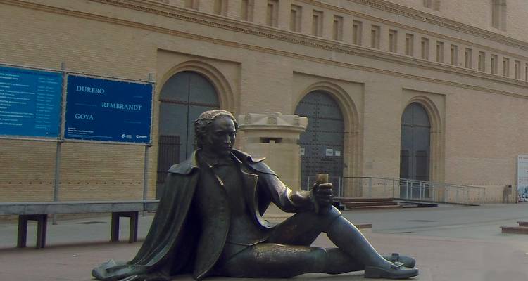 Estatua de bronce de un hombre sentado frente a un edificio grande.