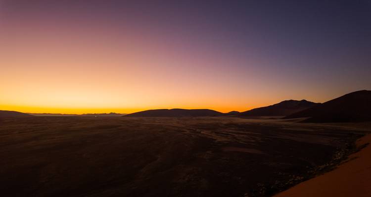 Weite Namib-Wüstenlandschaft bei Sonnenaufgang mit leuchtendem orangefarbenem Horizont und dunklen Dünen im Schatten.
