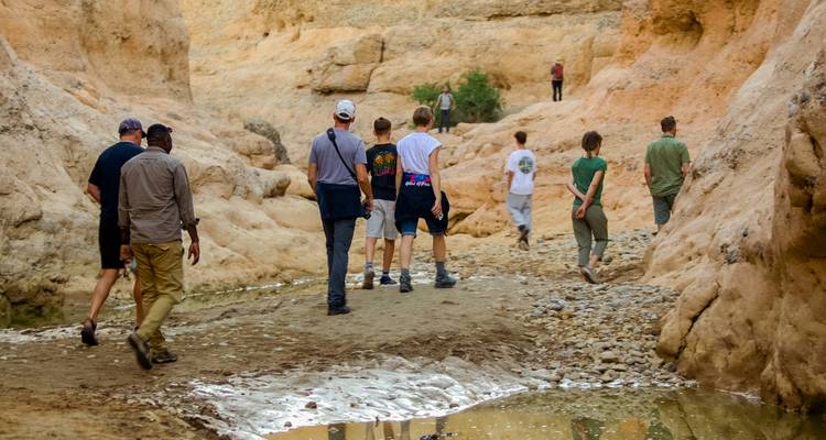 Reisende gehen durch einen engen, trockenen Canyon mit Pfützen, die ockerfarbene Wände widerspiegeln.