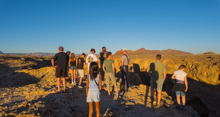 Eine Gruppe von Touristen versammelt sich am Rand einer sonnenbeschienenen Wüstendoline mit niedrigen Bergen in der Ferne.