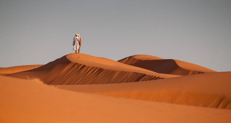 Silhouette d'une personne en tenue traditionnelle marchant sur des dunes de sable au coucher du soleil.