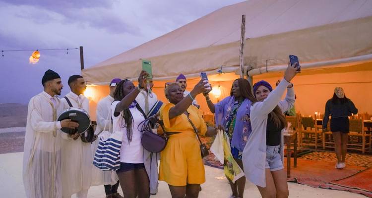 Glimlachende reizigers die selfies maken buiten een grote woestijntent bij schemering terwijl muzikanten in de buurt op trommels spelen