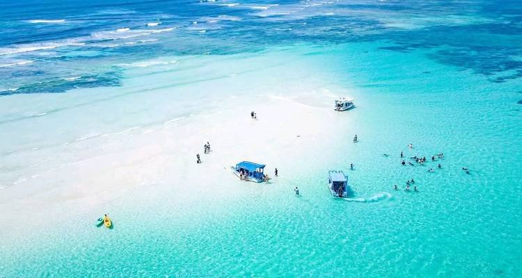 Vue aérienne d'une plage de sable blanc avec des bateaux et des personnes dans une eau turquoise.