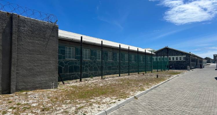 Buitenkant van een gevangenenblok met prikkeldraad op Robben Island onder een strakblauwe hemel.