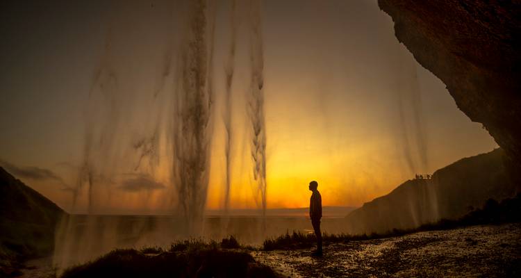 Silueta de una persona caminando detrás de una cascada al atardecer.