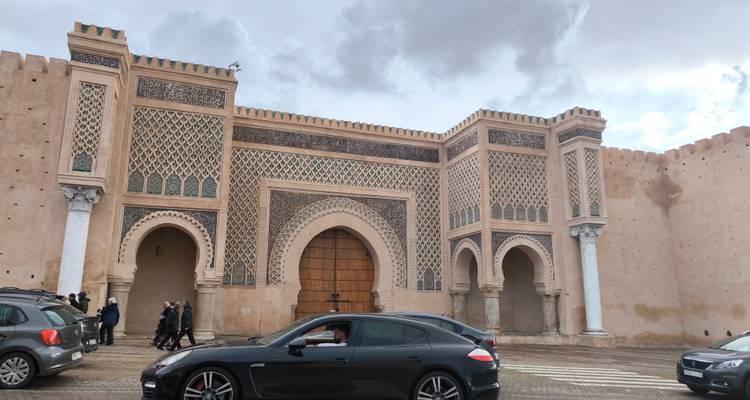 Een historische poort met ingewikkelde ontwerpen, voertuigen die voorbijrijden, en verschillende mensen in de omgeving.
