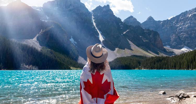Person wrapped in Canadian flag looking at mountain and lake landscape.