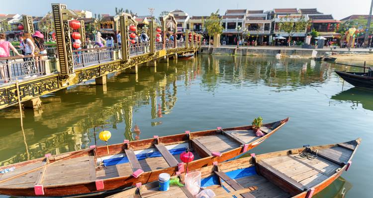Touristen überqueren eine geschmückte Brücke über einen kleinen Kanal mit Holzbooten in Hoi An.