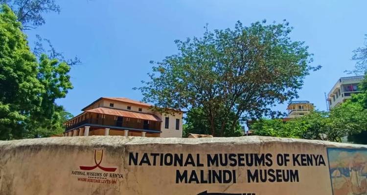 Panneau des Musées nationaux du Kenya avec des arbres et des bâtiments en arrière-plan sous un ciel bleu dégagé.