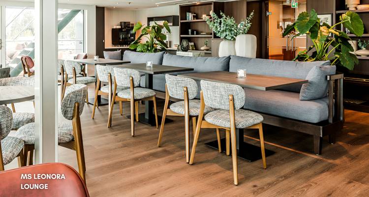 Loungebereich im Inneren eines Kreuzfahrtschiffs mit Tischen, Stühlen und Pflanzen.