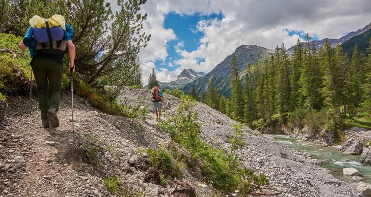 Zwei Wanderer erkunden einen Bergpfad mit Bäumen und einem Fluss.