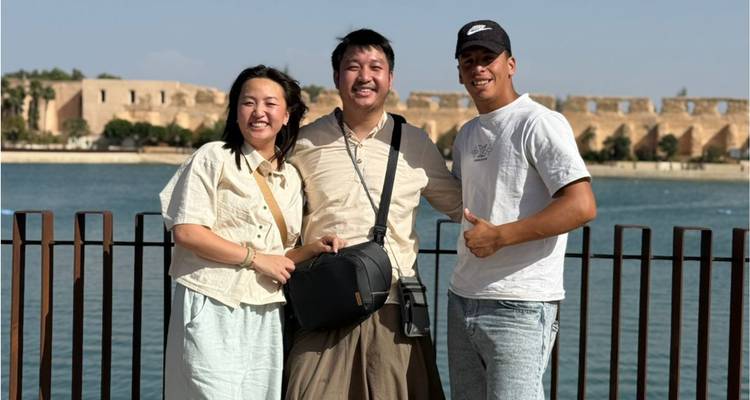 Trois voyageurs souriants à côté d'une balustrade au bord de l'eau avec les murs historiques de Rabat de l'autre côté du fleuve.