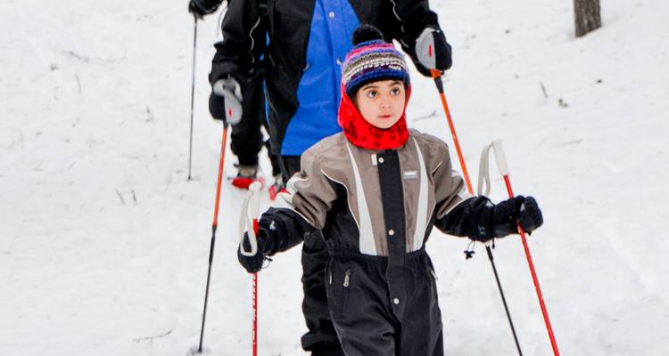 Ein Kind führt beim Skilanglauf an, während Erwachsene auf einer verschneiten Piste folgen.